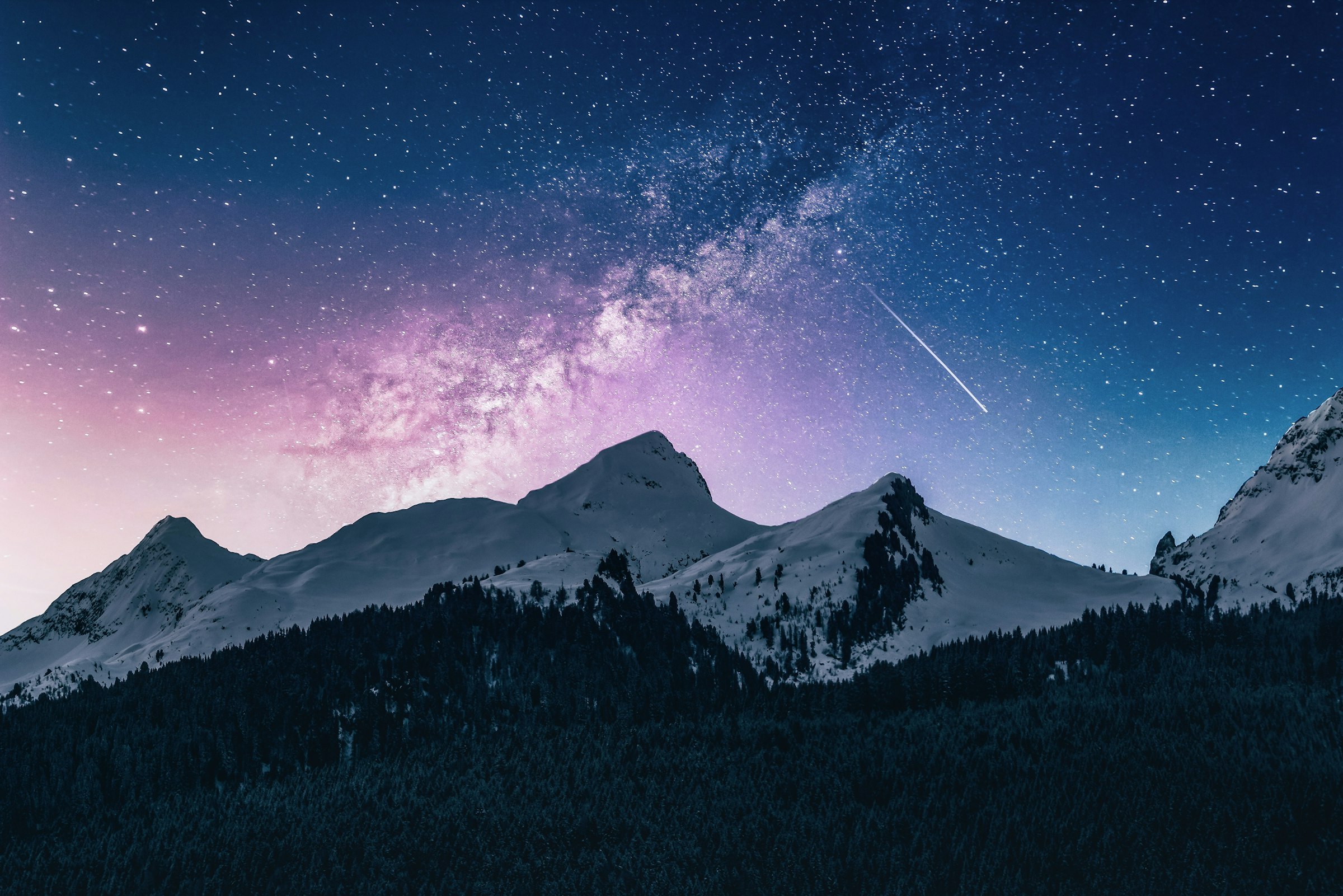 A sky full of stars over a snow-capped mountain and evergreen forest, warm light on the horizon